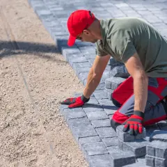 Pracownik w czerwonej czapce układa kostkę brukową. Zastanawiasz się, ile kosztuje ułożenie kostki brukowej? To zależy od wielu czynników.