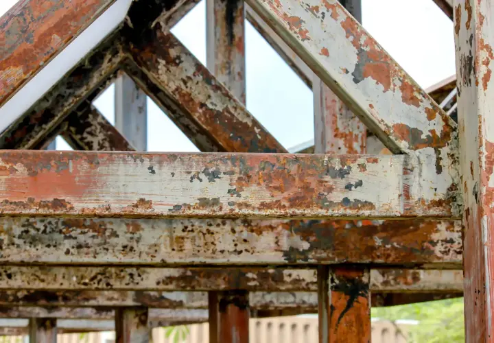 Metalowa konstrukcja z widocznymi śladami rdzy i łuszczącą się farbą. Wygląda na starą i zaniedbaną, jakby stała tam od lat.