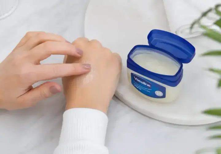 Dłonie rozsmarowują wazelinę techniczną na skórze, która staje się nawilżona i gładka. Słoiczek z produktem stoi obok.