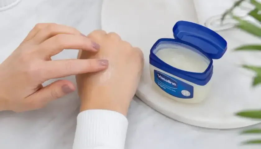 Dłonie rozsmarowują wazelinę techniczną na skórze, która staje się nawilżona i gładka. Słoiczek z produktem stoi obok.