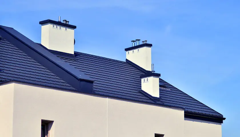 Nowoczesny dach bezokapowy z ciemnogranatową dachówką i białymi kominami, pod błękitnym niebem.