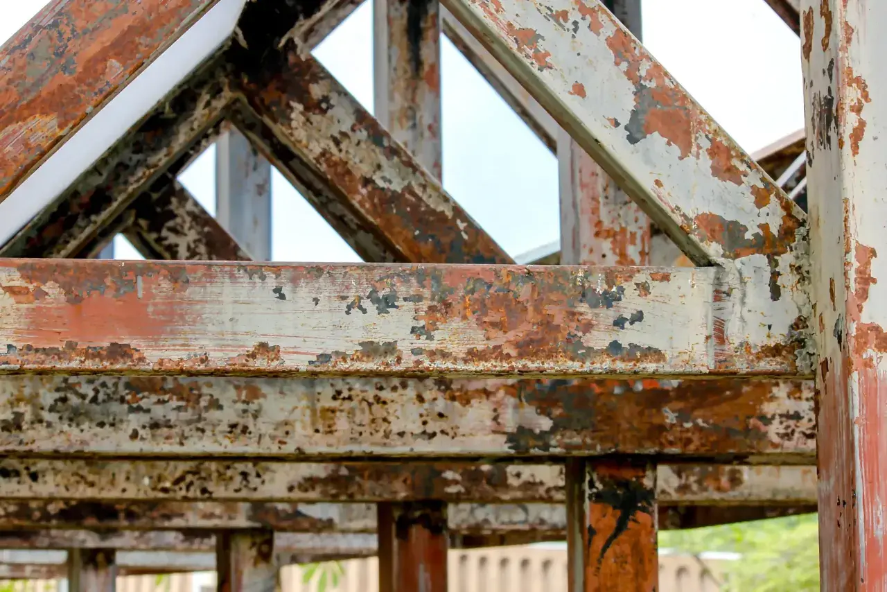 Metalowa konstrukcja z widocznymi śladami rdzy i łuszczącą się farbą. Wygląda na starą i zaniedbaną, jakby stała tam od lat.